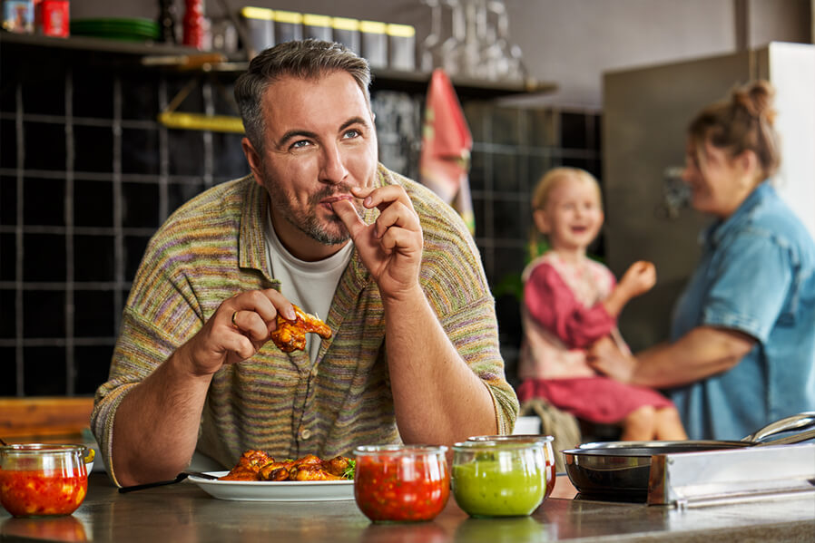 Familienvater genießt Chicken-Wings in der Küche und schleckt sich Finger ab.
